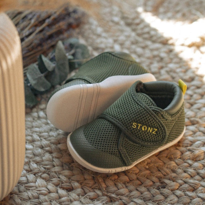Stonz Cruiser Original Baby Shoes Cypress green mesh with Velcro strap, resting on a woven rug beside a suitcase.