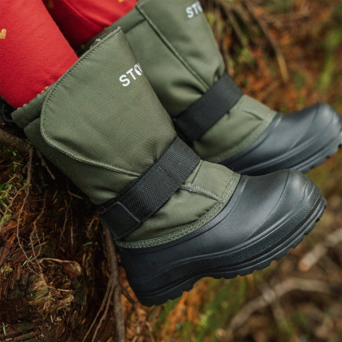 Stonz Trek Kid Snow Boots Cypress in green with Velcro straps secured, rugged black outsoles gripping a log.