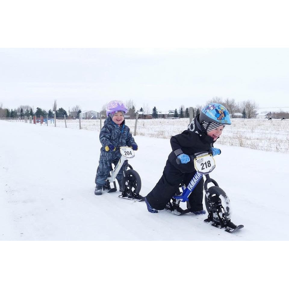 Strider 12 Snow Skis set; two kids in blue and purple outerwear riding on snow.