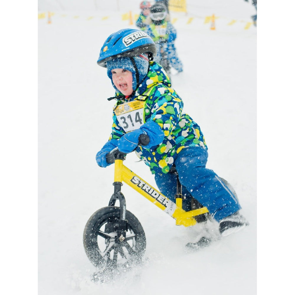 Strider 12 Snow Skis set; child in blue jacket and helmet rides a yellow STRIDER on a snowy slope.