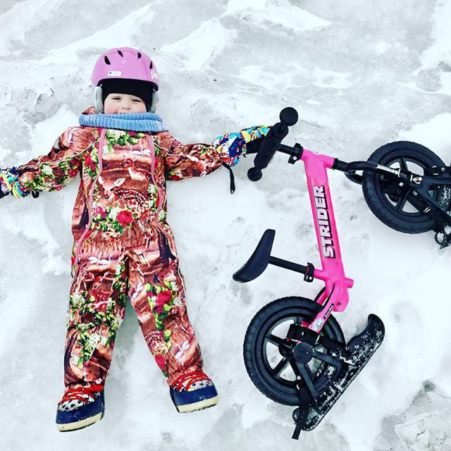 Strider 12 Snow Skis set for kids with pink frame; child in colorful snowsuit and pink helmet