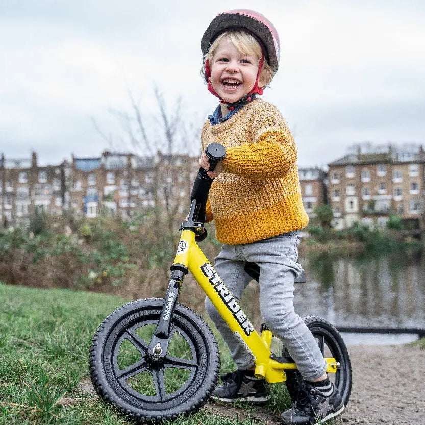 STRIDER 12 Sport balance bike in yellow, child confidently rides on a grassy path wearing a helmet.
