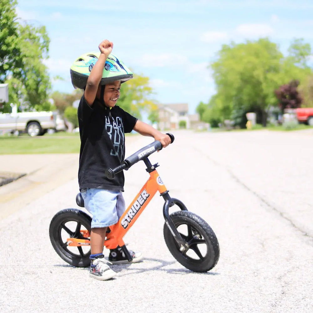 Strider 12 Sport balance bike, orange frame with black handlebars, child raises fist wearing helmet.
