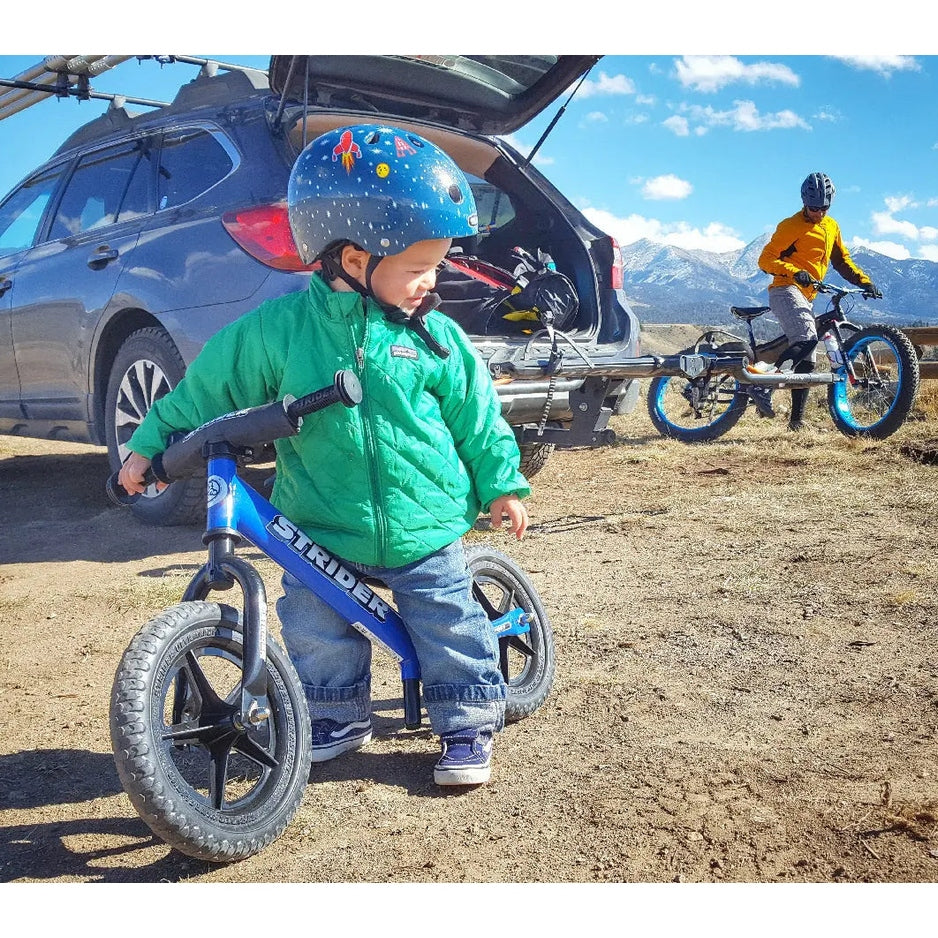 Strider 12 Sport balance bike; blue frame helps a toddler in a green jacket balance on a dirt lot.
