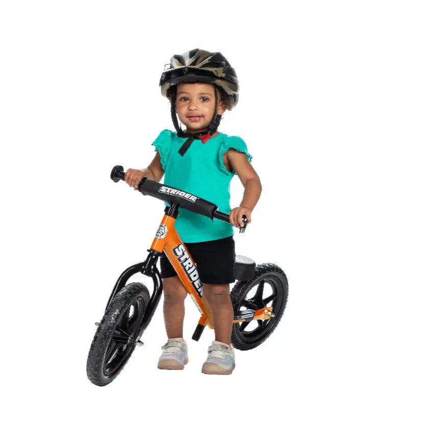 Strider 12 Sport balance bike, orange frame; toddler in teal shirt wearing a helmet pushes along.