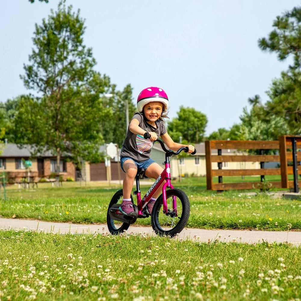 Strider 14X Classic balance bike in pink; a child rides along a park path wearing a pink helmet.