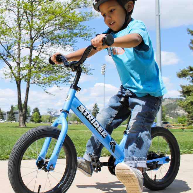 Strider 14X Classic Balance Bike in blue; a kid rides outdoors on a sunny day, gripping handlebars with helmet on.