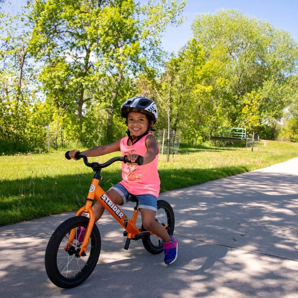 Strider 14X Classic Balance Bike with orange frame, ridden by a smiling child in a pink top on a sunny park path.