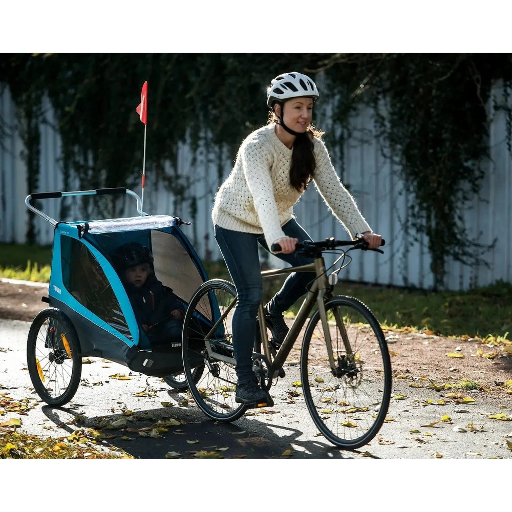 Thule Coaster Xt bike trailer in blue and gray, child seated inside while a cyclist parent rides along.