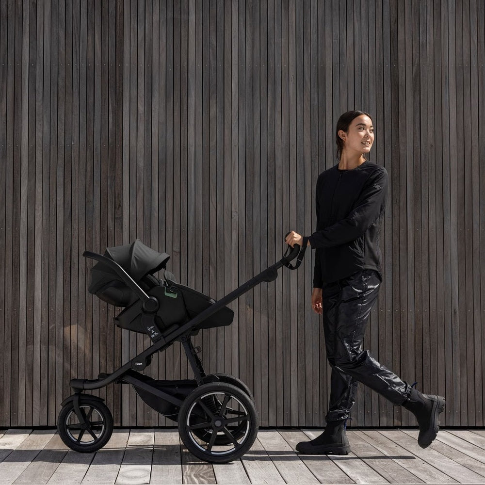 THULE Urban Glide 3 stroller in black, pushed by a woman along a wooden wall with large wheels.