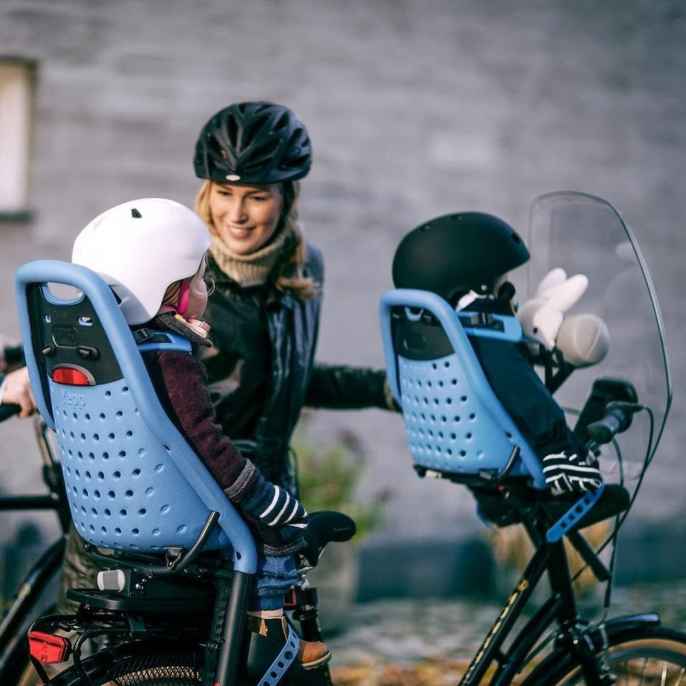 Thule Yepp Maxi rack-mounted child bike seats on blue bikes; parent helps helmeted child buckle in.