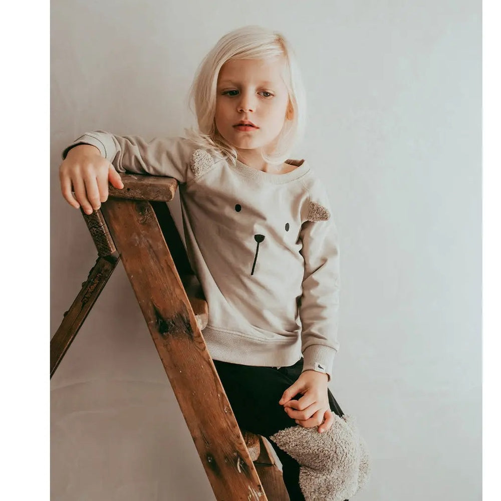 Turtledove London 3D Character Sweatshirt, pumice beige, child leans on a wooden ladder showing bear embroidery.