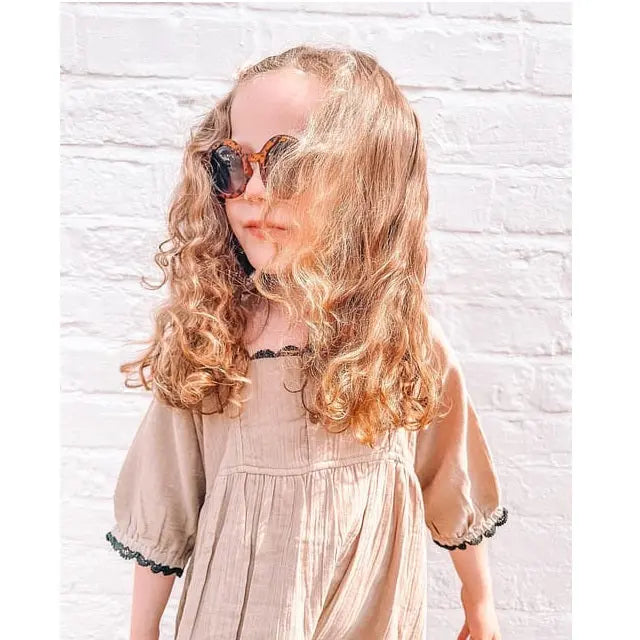 Turtledove London Herb Gauze Dress on a toddler in beige puff sleeves with lace trim, posing in sunglasses.