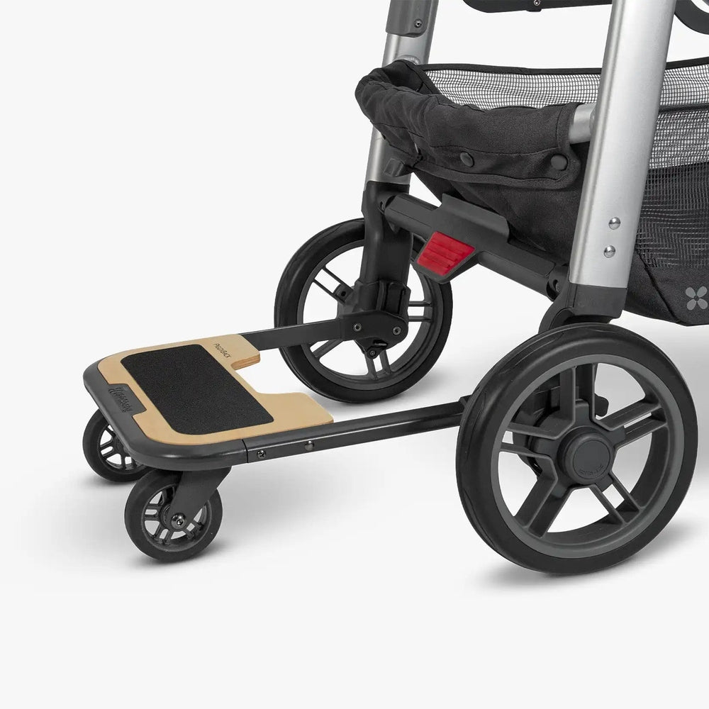 UPPAbaby Cruz PiggyBack board mounted on the rear of a stroller, close-up side view showing attachment point, standing platform, and small front wheels.