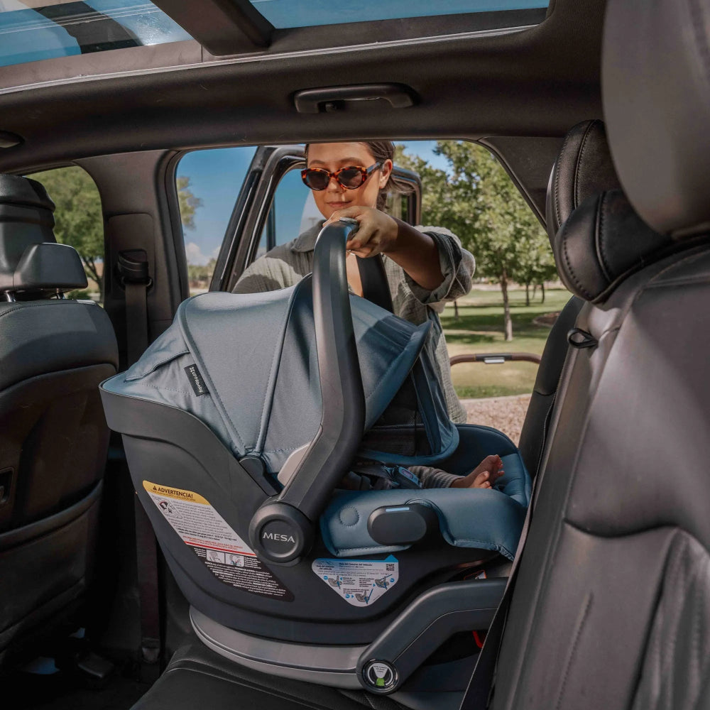 UPPABABY Mesa V3 Infant Car Seat in blue-gray being loaded into a car by a parent, showing the easy-grip handle.