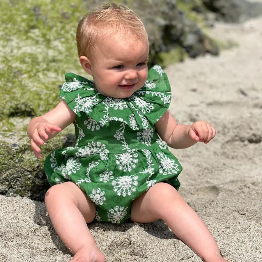 Vignette Maya Bubble Green Eyelet baby romper; green with white floral eyelet, baby sits on beach.