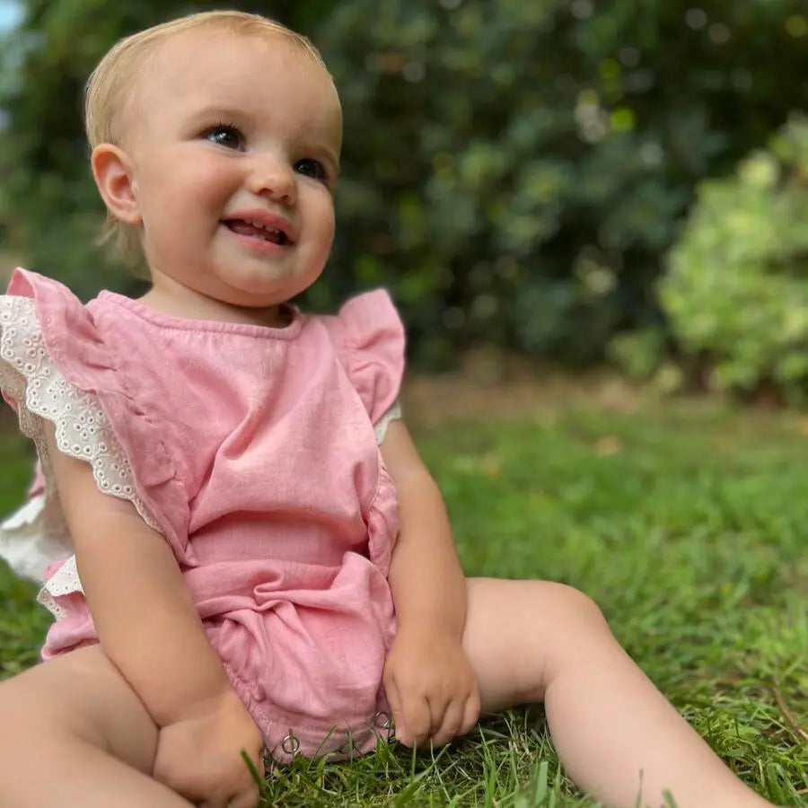 Vignette Megan Bubble Pink baby romper; pink fabric with lace-trim sleeves, a smiling toddler sitting on grass outdoors.