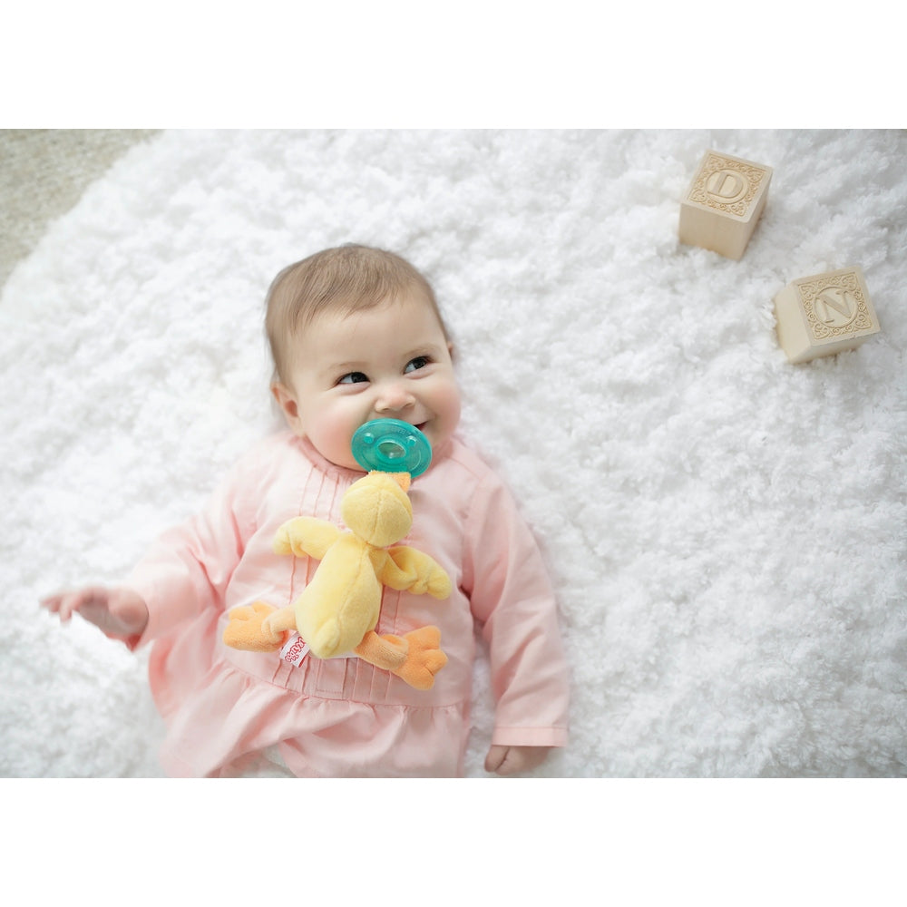 WubbaNub Infant Plush Toy Pacifier Orange Duck by Aurora; baby smiles as teal pacifier is attached to the yellow plush duck on a fluffy rug.