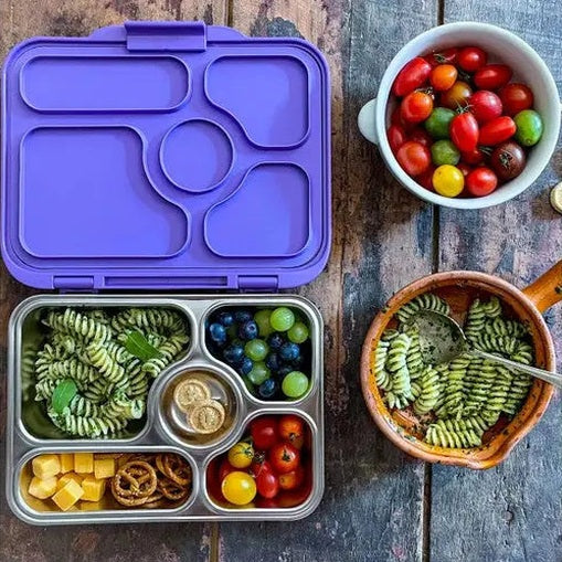 Yumbox Presto lunch box set with purple lid, open to reveal divided metal tray holding grapes, blueberries, cheese, pasta, and tomatoes.